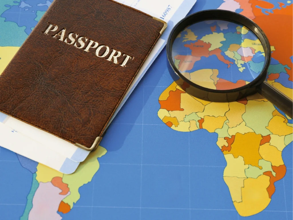 Ein brauner Reisepass mit herausstehendem Dokument und eine Lupe liegen auf einer bunten Weltkarte mit Fokus auf den afrikanischen und europäischen Kontinent.